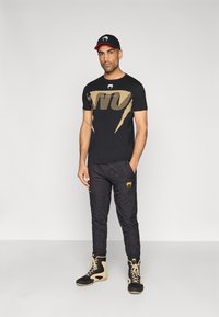 Zwart t-shirt met gouden grafisch patroon, zwarte broek met patroon en zwarte laarzen met beige accenten. Hoed met logo en rode rand.