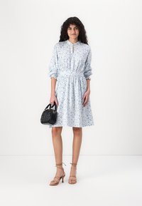 Vestido floral azul claro con un cuello alto, cintura elástica y mangas abullonadas. Combinado con un bolso de mano tejido negro y tacones beige con tiras.