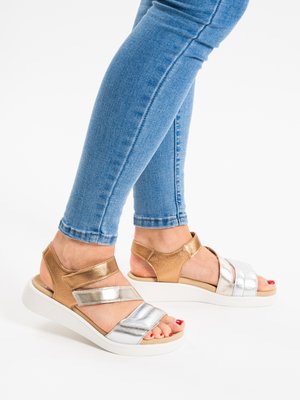 Fødder iført metaliske roseguld- og sølvsandaler med hvide såler, kombineret med tætsiddende blå jeans mod en hvid baggrund.