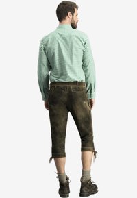 Grün kariertes Hemd kombiniert mit braunen Lederhosen, die Seitentaschen und Träger haben, getragen mit grauen Socken und braunen Schuhen.