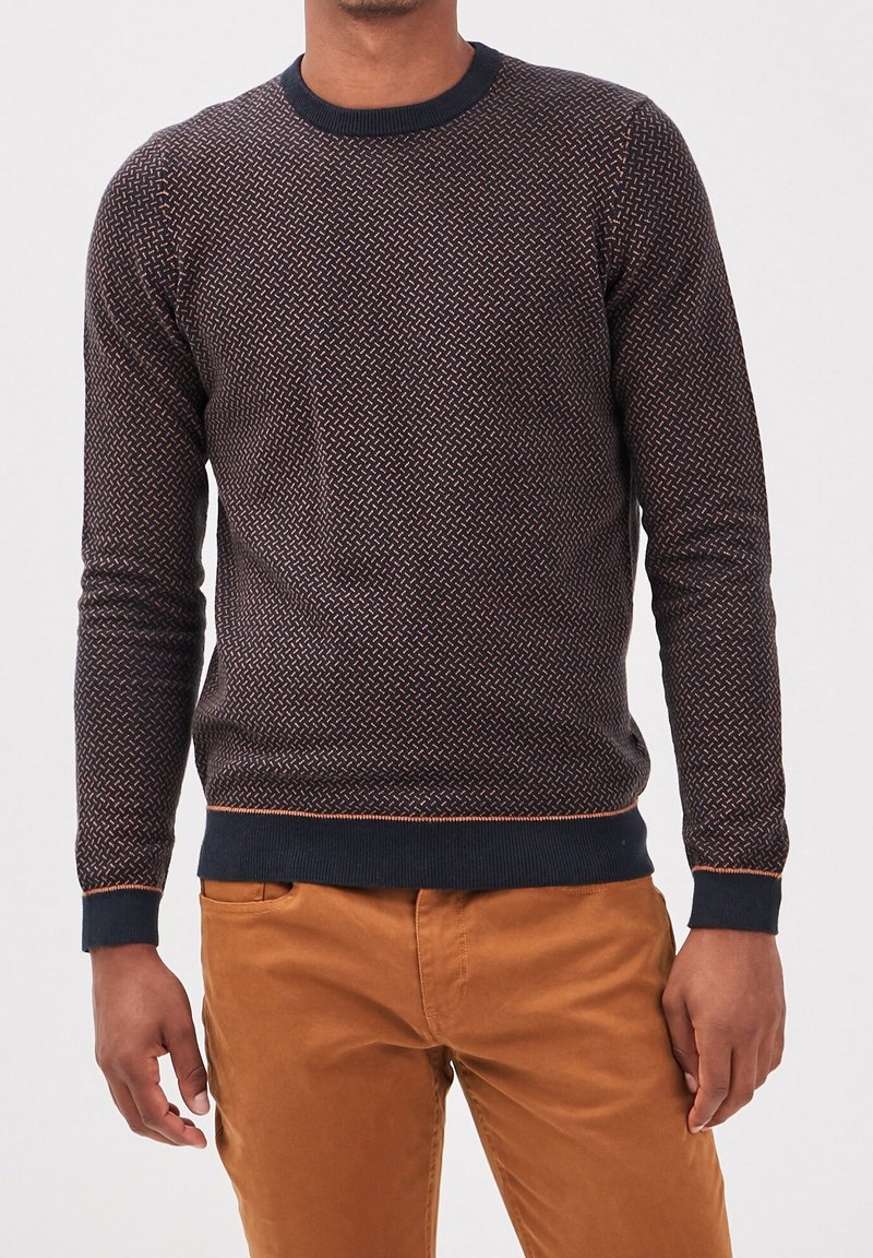 Hombre vistiendo un suéter de manga larga azul marino con un patrón y acentos naranjas, y pantalones color mostaza, de pie frente a un fondo liso.