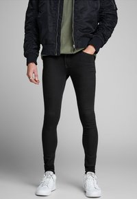 Pantalones vaqueros ajustados negros, confeccionados en denim, con un diseño elegante y una textura suave, combinados con una camisa verde y una chaqueta bomber negra.