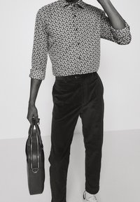 Zwart-wit overhemd met patroon en kraag, gecombineerd met donkere corduroy broek. Houdt een gestructureerde zwarte tas vast, met een gladde witte sneaker zichtbaar.