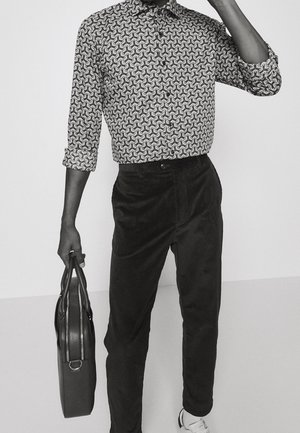 Czarna i biała koszula w wzory z kołnierzykiem, zestawiona z ciemnymi sztruksowymi spodniami. Trzyma teksturowaną czarną torbę, widoczny jest gładki biały sneaker.