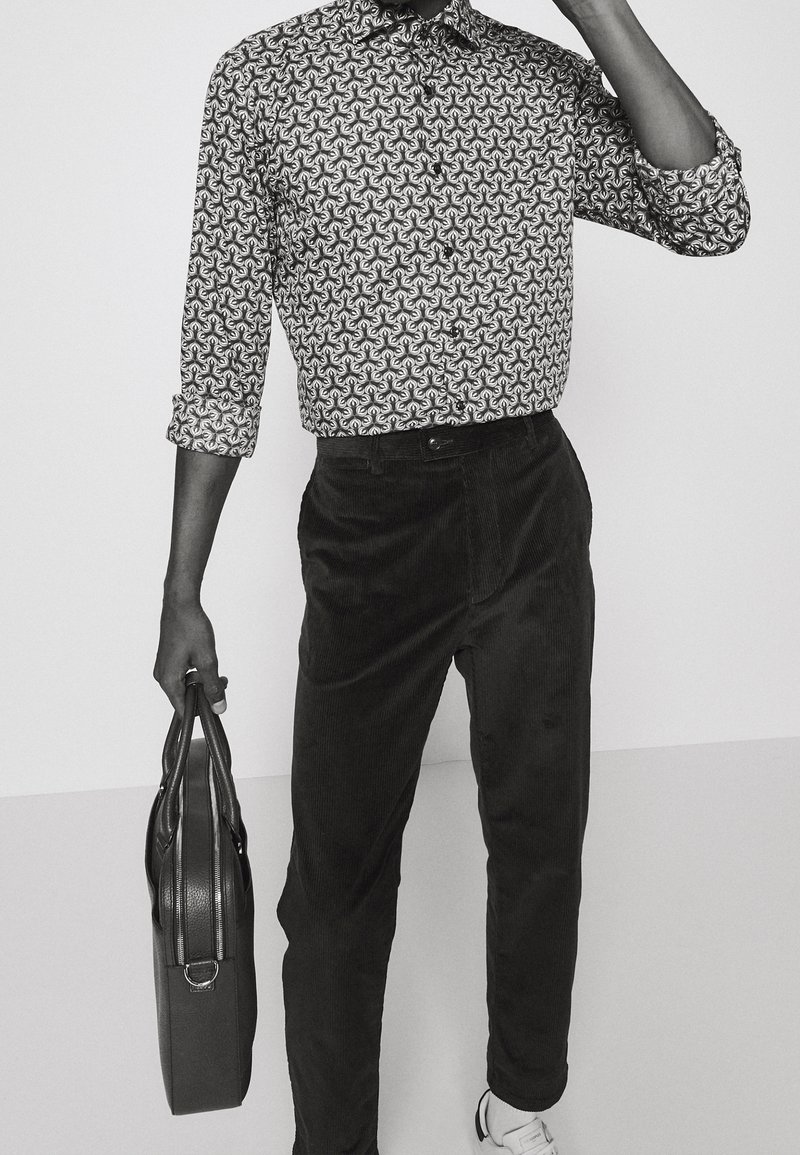 Zwart-wit overhemd met patroon en kraag, gecombineerd met donkere corduroy broek. Houdt een gestructureerde zwarte tas vast, met een gladde witte sneaker zichtbaar.