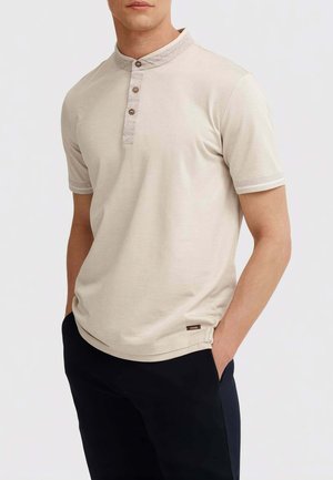Uomo che indossa una maglia henley beige a maniche corte con patta a bottoni a contrasto e pantaloni neri, con le mani nelle tasche, su sfondo semplice.