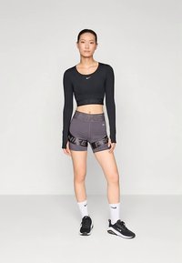 Melnas garas piedurknes apgrieztas blūze un pelēki šorti ar uzrakstu "NIKE PRO", apvienoti ar melnām sporta kurpēm un baltām zeķēm, stāvot uz neitrāla fona.