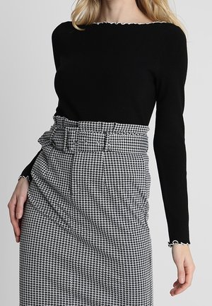 Femme portant un haut noir cintré à manches longues et une jupe taille haute à motif pied-de-poule noir et blanc avec ceinture assortie.