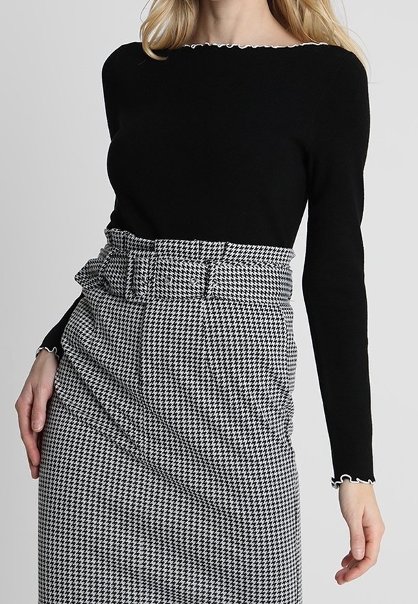 Femme portant un haut noir cintré à manches longues et une jupe taille haute à motif pied-de-poule noir et blanc avec ceinture assortie.