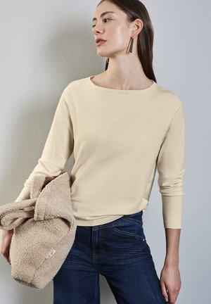 Maglietta beige a maniche lunghe con collo rotondo, abbinata a jeans blu scuro. Il modello tiene un capo beige morbido, simile al pile.