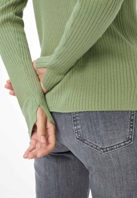 Pull à manches longues côtelé de couleur vert clair, avec des poignets fendus et une coupe ajustée. Associé à un jean gris délavé moyen, mettant en valeur le détail des poches arrière.