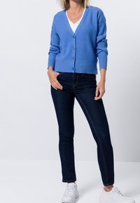 Světle modrý žebrovaný kardigan s dlouhými rukávy, výstřihem do V a knoflíky vpředu, kombinovaný s tmavými džínami a bílými teniskami.