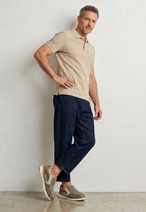 Mężczyzna stoi z ręką na biodrze, ubrany w beżową koszulkę polo, ciemnoniebieskie krótkie spodnie i szare sneakersy na wsuwkę, na tle jednolitej ściany.