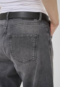 Jeans en denim gris avec une coupe droite, dotés de poches arrière et d'une ceinture en cuir noir. Le tissu a une texture délavée et des coutures subtiles.
