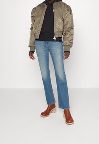 Chaqueta bomber verde oliva con puños acanalados, sudadera negra debajo, jeans rectos de mezclilla azul claro y zuecos de tacón marrón con suelas de madera.