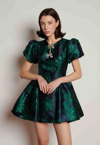 Σύντομο φόρεμα από σατέν με βάση σε σκούρο ναυτικό μπλε χρώμα, πράσινα λουλουδάτα σχέδια, φουσκωτά μανίκια και ασημένιες φιόγκους στο μπροστινό μέρος.