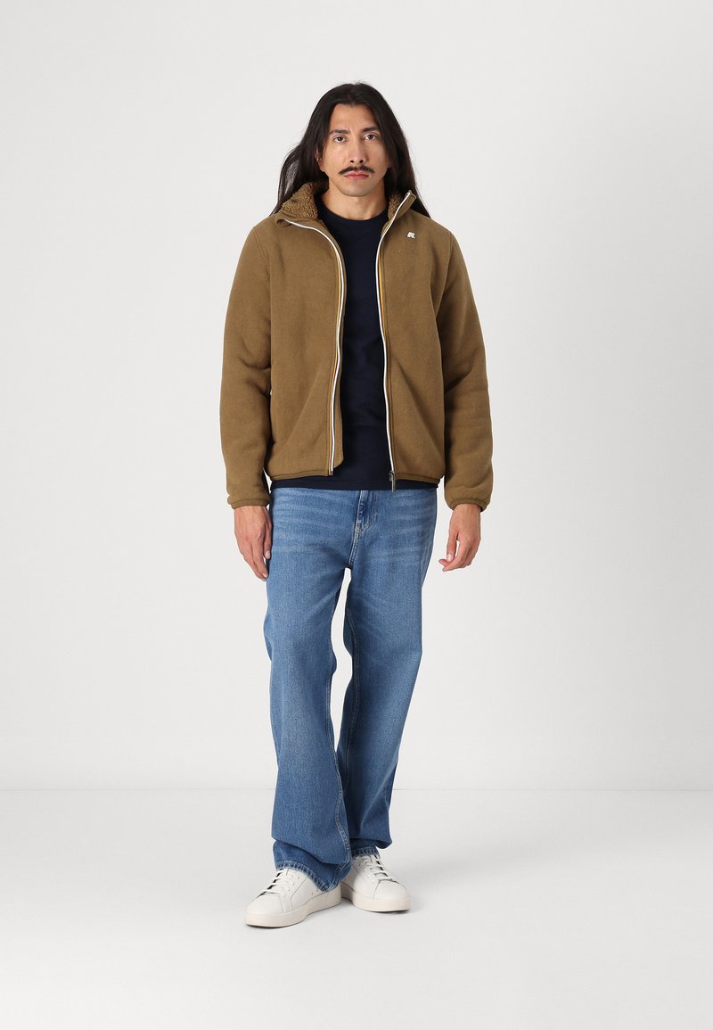 Braune Fleecejacke mit Reißverschluss und leichten Akzenten, getragen über einem dunkelblauen Hemd, kombiniert mit hellblauen Jeans und weißen Sneakers.