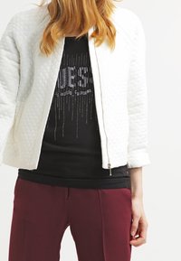 Veste bomber matelassée blanche avec une fermeture éclair, des poignets ajustés et un design légèrement court, portée sur un t-shirt graphique noir et un pantalon bordeaux.