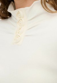 Biały sweter z wysokim kołnierzem, o prążkowanej fakturze, z dekoracyjnymi guzikami z tkaniny w kształcie róż wzdłuż listwy guzikowej.