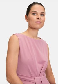 En rosa ärmlös klänning med rund halsringning och plisserad design med midjeband. Tillverkad av mjukt, texturerat tyg.