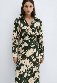 Cvjetna haljina na preklop u tamnozelenoj boji, s velikim breskvinim cvjetovima, dugim rukavima, kragnom i detaljem s vezanjem u struku. Glatka tekstura tkanine.