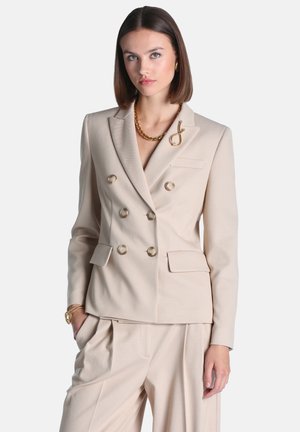 Kvinde med glat brunt hår iført en beige dobbeltradet blazer og matchende bukser med guldkæde og armbånd.