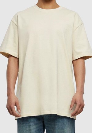 Osoba ubrana w luźny, beżowy t-shirt z krótkim rękawem i niebieskie dżinsy, stojąca na jednolitym jasnoszarym tle.