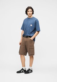 Νέος άνδρας όρθιος με τα χέρια στις τσέπες, φορώντας μπλε μπλουζάκι, καφέ σορτς cargo, λευκές κάλτσες και μαύρα αθλητικά παπούτσια σε απλό φόντο.