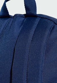 Zaino in tessuto blu navy in primo piano, con materiale testurizzato e cuciture visibili, spallacci imbottiti e una maniglia superiore rinforzata.