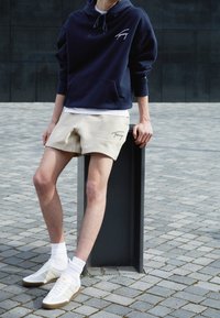 Mörkblå huvtröja med vitt logotyp, beige shorts med logotyp, vita strumpor och vita sneakers med guld accenter, mot en grå belagd bakgrund.