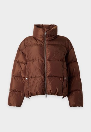 Femme aux cheveux blonds lisses portant une veste matelassée marron, un pantalon évasé noir et des talons pointus noirs, debout devant un fond uni.