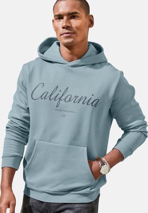 Man wearing light blue hoodie with "California State College" text, one hand in front pocket, and wristwatch on the other wrist.