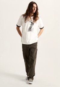 White cotton t-shirt with black ribbed cuffs featuring a graphic design of a serpent with flames. Paired with loose-fit olive green pants.