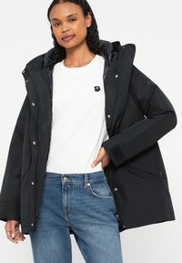 Μαύρο αδιάβροχο μπουφάν με κουκούλα, με κουμπώματα, και επενδυμένο εσωτερικό. Φορεμένο πάνω από ένα λευκό μπλουζάκι με ένα μικρό λογότυπο μαύρου λουλουδιού.