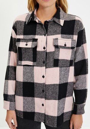 Frau trägt ein schwarz-pink kariertes Hemd mit Knöpfen, zwei aufgesetzten Taschen vorne und langen Ärmeln.