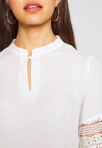Blouse blanche avec un col montant, petite fermeture à fente sur le devant et des détails en dentelle sur les manches courtes. Texture lisse et tissu léger.