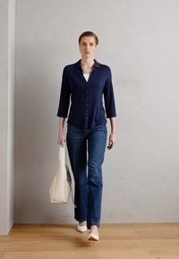 Marineblå skjorte med knapper, trekvartlange ermer, kombinert med blå jeans. Modellen holder en kremfarget toteveske og har på seg hvite hæler.