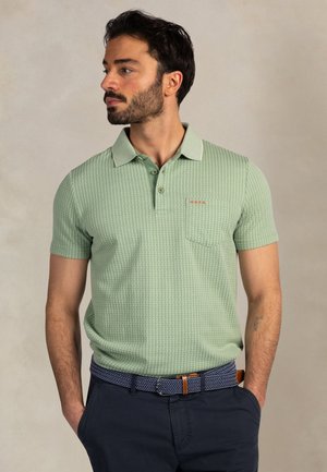 Man met baard, gekleed in een lichtgroen poloshirt met patroon en donkerblauwe broek met een blauwe gevlochten riem, kijkt naar links.
