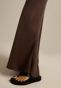 Długa, zwiewna brązowa sukienka z lekkim rozcięciem po boku, zestawiona z czarnymi klapkami, które mają teksturowaną podeszwę i szeroki pasek.