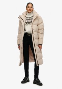Beige lange pufferjas met hoge kraag, voorkant met rits, en zijzakken, gedragen over een gebreid patroontrui en zwarte jeans met veterschoenen.