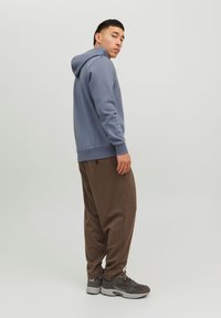 Niebieska bluza z kapturem o miękkiej fakturze, brązowe spodnie tapered i szare sneakersy, widok z boku na jasnym tle.