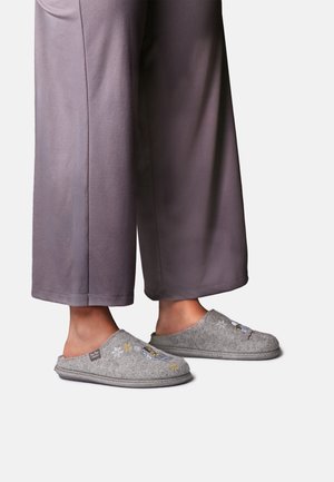 Fødder iført grå filt-slip-on tøfler med små broderede motiver, kombineret med løse grå bukser mod en ensfarvet baggrund.
