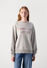 Šedá mikina s ribovaným kulatým výstřihem, dlouhými rukávy a vínově červeným potiskem "CONNECTICUT GANT 1949" na přední straně. Nosí se s modrými džíny.