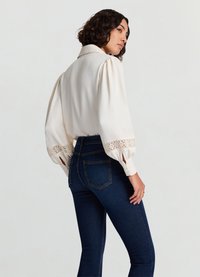 Blouse crème à manches bouffantes, détails en dentelle sur les poignets ; associée à un jean slim bleu foncé. Tissu lisse, design structuré et texture subtile.
