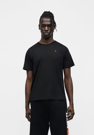 Uomo in piedi, in posizione eretta, che indossa una maglietta nera semplice e pantaloncini neri con un logo bianco e rosso sulla gamba sinistra, su sfondo bianco.