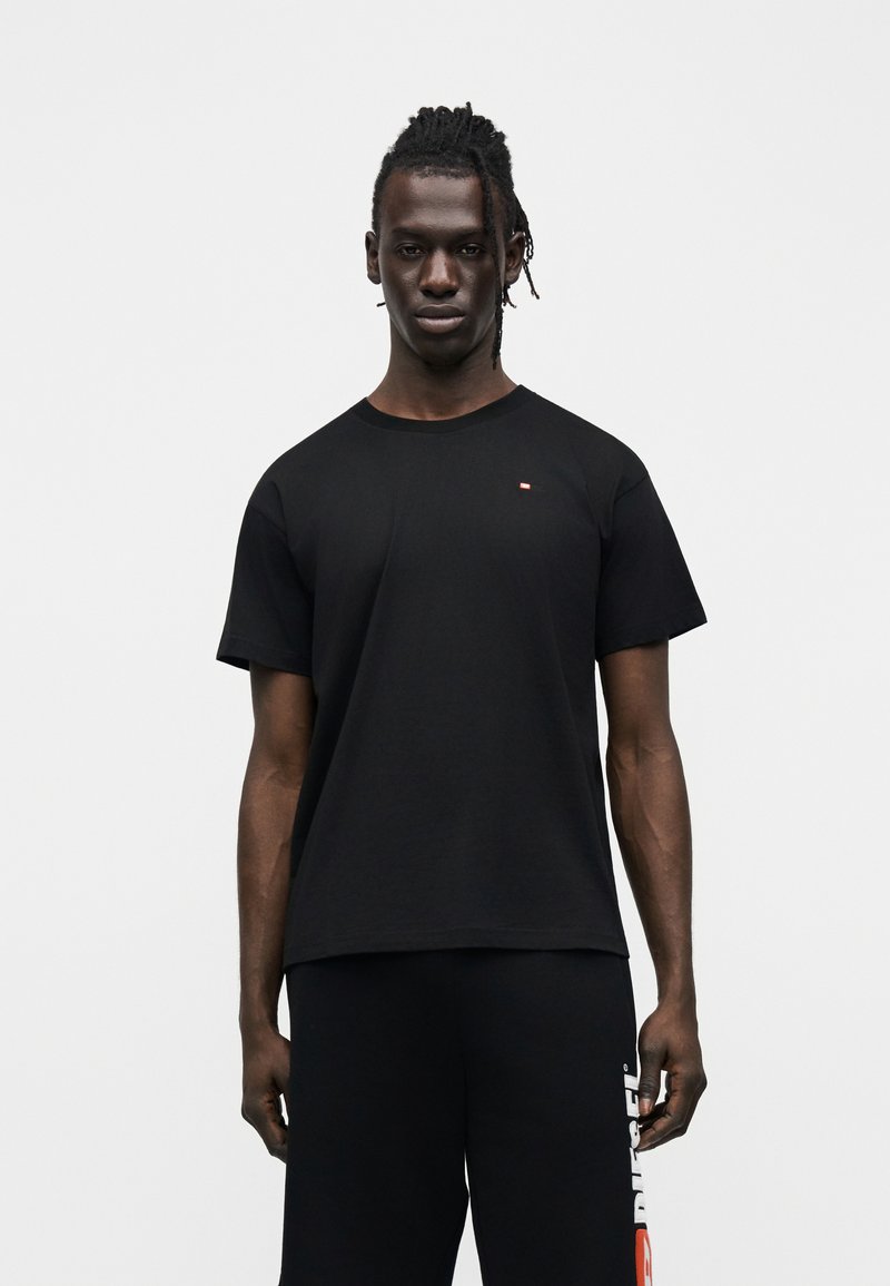 Άνδρας σε όρθια στάση, φοράει μονόχρωμη μαύρη μπλούζα και μαύρο σορτς με λευκό και κόκκινο λογότυπο στο αριστερό πόδι, μπροστά σε λευκό φόντο.