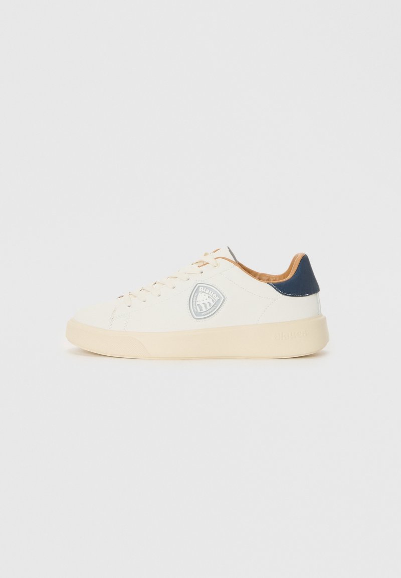 Baskets basses blanches avec semelle beige, languette de talon bleu marine, intérieur beige, lacets et un patch logo circulaire sur le côté.