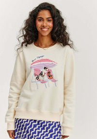 Krémová mikina s tištěným designem znázorňujícím psa a osobu na pruhovaných lehátkách pod slunečníkem. Modrá sukně s vlnitým vzorem dole.