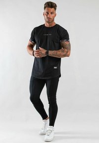 Svart t-shirt med kort ärm och dekorativa mönstrade manschetter, kombinerad med åtsittande svarta byxor och vita sneakers. Grafisk text i mitten av t-shirten.