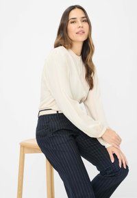Lys beige bluse med høj hals og pufærmer, parret med mørkeblå stribede bukser, siddende på en træstol.